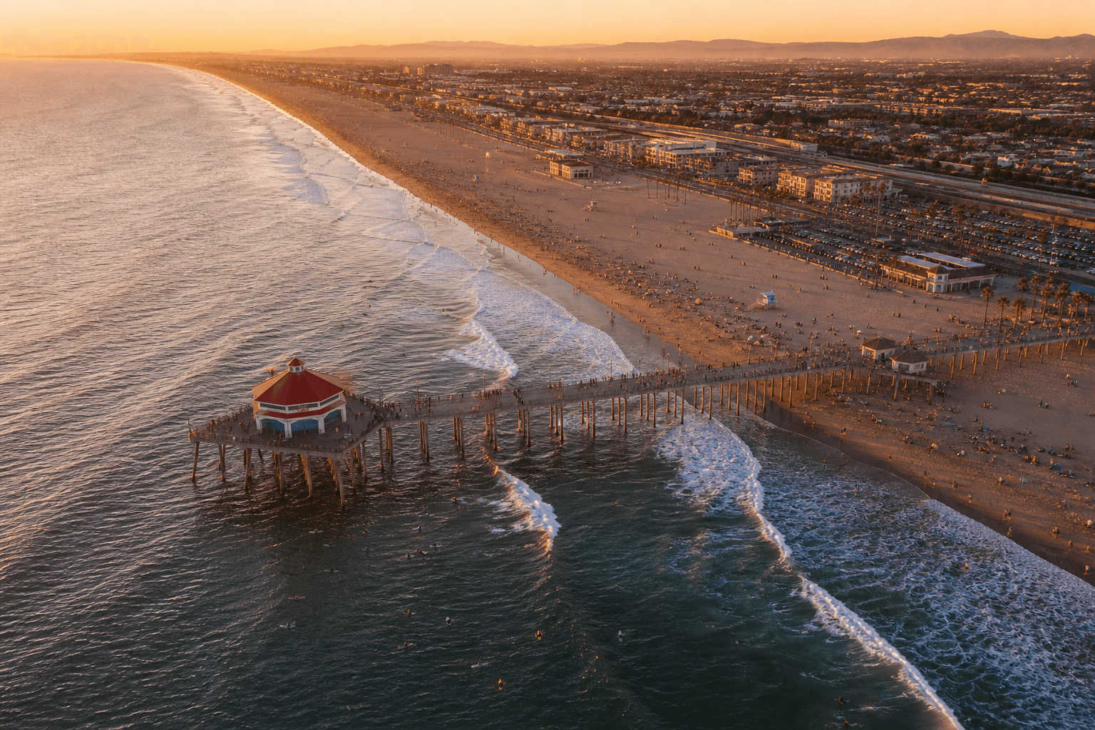 Aerial golden-hour view of Huntington Beach California pier stretching over the Pacific Ocean with surfers and the long sandy coastline